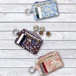 May include: Fabric mini wallets in blue floral, purple floral, and pink floral patterns. Each wallet has a zipper closure, a key ring, and card slots. Finished size is approximately 3" x 4 3/4". The text reads "Fabric Mini Wallet".
