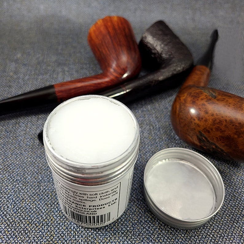 Może przedstawiać: Mały srebrny pojemnik z białą substancją w środku. Pojemnik jest oznaczony etykietą "Pipe Products Ltd." i ma kod kreskowy. W tle znajdują się trzy drewniane fajki.