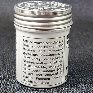 Może przedstawiać: Biały słoik ze srebrną pokrywką. Słoik zawiera wosk do polerowania. Etykieta na słoiku głosi: "Rafinowane woski zmieszane według formuły stosowanej przez British Museum i specjalist&oacute;w od renowacji na całym świecie, aby ożywić i chronić cenne meble, sk&oacute;rę, obrazy, metale, marmur, kość słoniową i wiele innych powierzchni, zar&oacute;wno w pomieszczeniach, jak i na zewnątrz. Odświeża kolory, nadaje delikatny połysk."