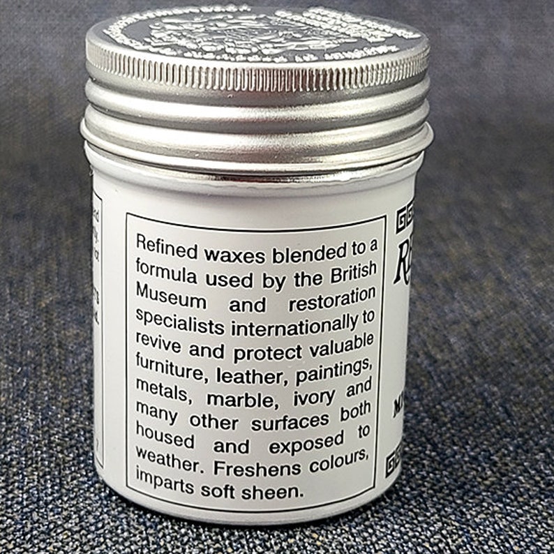 Może przedstawiać: Biały słoik ze srebrną pokrywką. Słoik zawiera wosk do polerowania. Etykieta na słoiku głosi: "Rafinowane woski zmieszane według formuły stosowanej przez British Museum i specjalist&oacute;w od renowacji na całym świecie, aby ożywić i chronić cenne meble, sk&oacute;rę, obrazy, metale, marmur, kość słoniową i wiele innych powierzchni, zar&oacute;wno w pomieszczeniach, jak i na zewnątrz. Odświeża kolory, nadaje delikatny połysk."