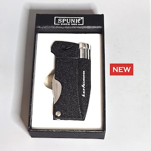 May include: A black textured lighter with a silver blade and a silver top, presented in a black box with a white interior. The box has the text "SPUNK SINCE 1989". A red "NEW" sticker is visible.