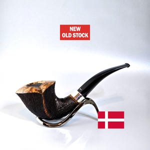 Peut inclure: Une pipe à tabac en bruyère avec un foyer foncé et texturé et un tuyau noir brillant. La pipe repose sur un support argenté, avec le texte "NEW OLD STOCK" dans un rectangle rouge au-dessus. Un drapeau danois est dans le coin inférieur droit.