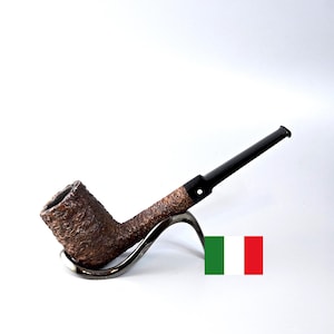 Puede incluir: Una pipa de tabaco marrón con un cuenco texturizado y una boquilla negra. La pipa descansa sobre un soporte plateado sobre un fondo blanco. Un accesorio clásico para fumadores.