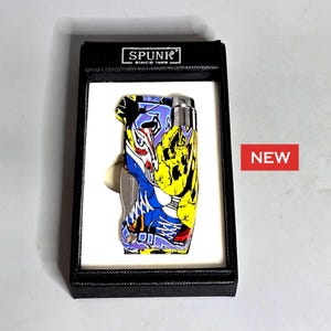 May include: A colorful, graffiti-style lighter with a black box. The lighter features a vibrant design with blue, yellow, purple, and red accents. The box has the text "SPUNK SINCE 1989" printed on it. A red "NEW" sticker is visible.
