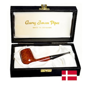 May include: A brown wooden tobacco pipe with a black stem resting inside a black rectangular presentation box lined with white satin fabric. The interior lid features the gold text Georg Jensen Pipes MADE IN DENMARK above the pipe.
