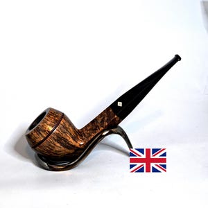 May include: A tobacco pipe with a polished brown wood grain bowl and a long, tapered black stem. The stem features a small white diamond-shaped inlay near the bowl. The pipe rests on a curved metal stand, accompanied by a small Union Jack flag icon.