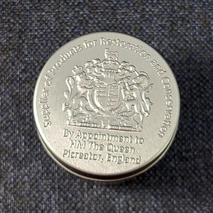 Może przedstawiać: Okrągłe, srebrne pudełko z napisem "Supplier of Products for Restoration and Conservation" oraz "By Appointment to HM The Queen Picreator, England". Pudełko zdobi herb kr&oacute;lewski z lwem i jednorożcem.