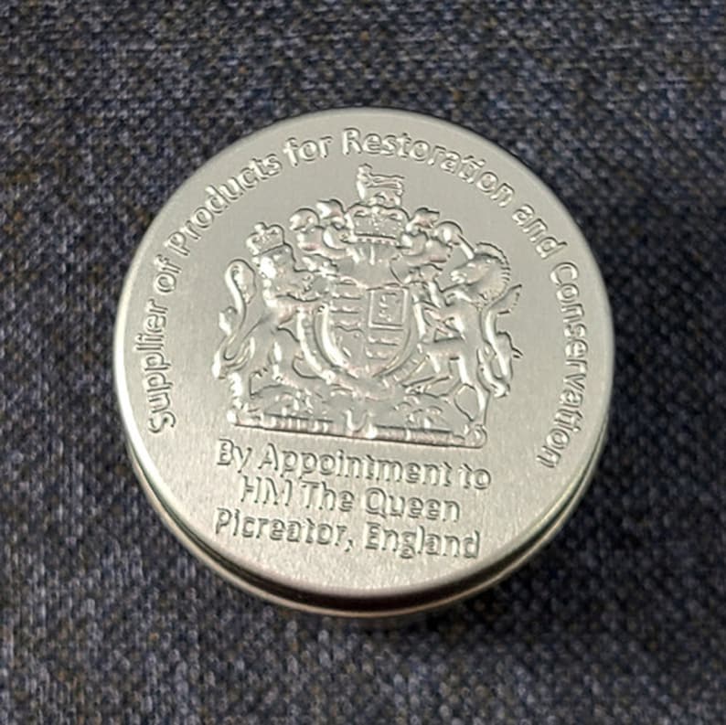 Może przedstawiać: Okrągłe, srebrne pudełko z napisem "Supplier of Products for Restoration and Conservation" oraz "By Appointment to HM The Queen Picreator, England". Pudełko zdobi herb kr&oacute;lewski z lwem i jednorożcem.