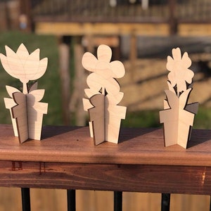 May include: Three wooden flower pot decorations with different flower designs. The flowers are cut out of wood and stand upright in the pots. The pots are also made of wood and have a simple design.