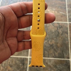 May include: Yellow silicone watch band with a sunflower design. The band features a repeating pattern of sunflowers and is designed for a smartwatch. The band is held in a hand.