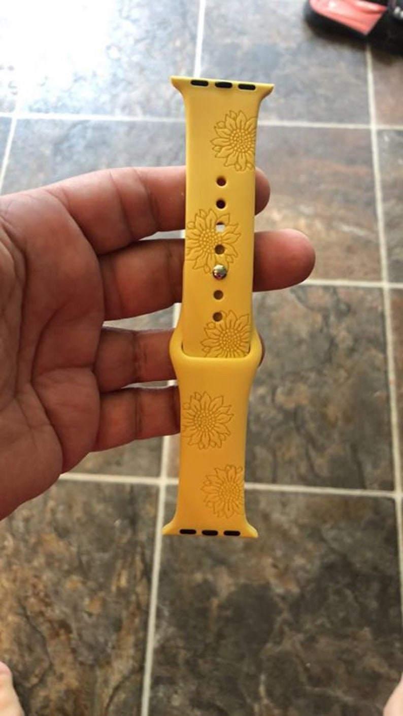 May include: Yellow silicone watch band with a sunflower design. The band features a repeating pattern of sunflowers and is designed for a smartwatch. The band is held in a hand.