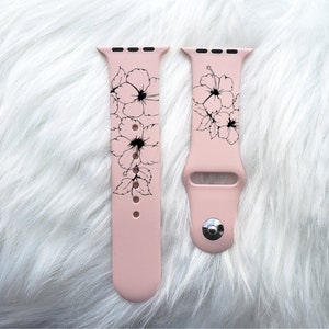 May include: A pink silicone Apple Watch band with a black floral design. The band is shown on a white fuzzy surface.