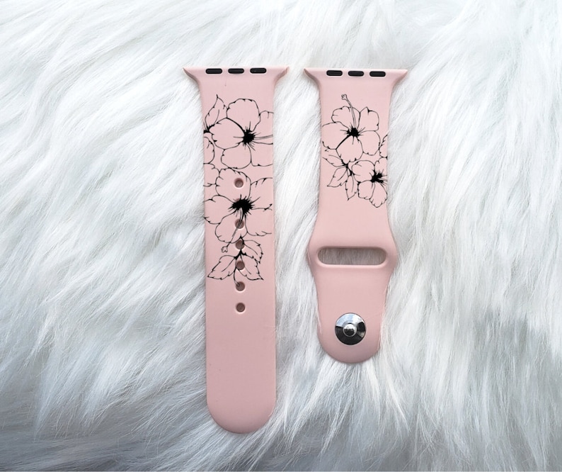 May include: A pink silicone Apple Watch band with a black floral design. The band is shown on a white fuzzy surface.
