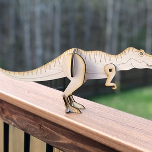 Op de afbeelding: Een houten T-Rex-dinosaurusspeelgoed met scharnierende poten en staart. De dinosaurus is lichtbruin en heeft een gedetailleerd ontwerp.