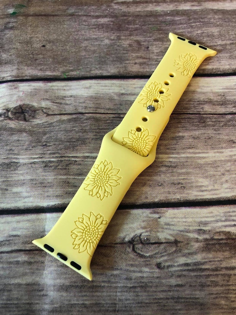May include: A light yellow Apple Watch band with a sunflower design. The band is made of a flexible material and features three embossed sunflower designs. The band is shown on a wooden surface.