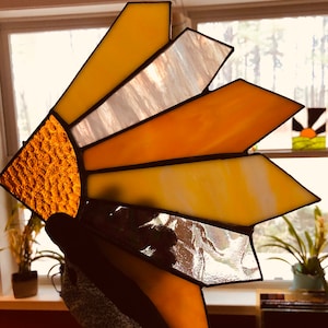 Puede incluir: Un atrapasoles de vidriera con un dise&ntilde;o de sol. El atrapasoles presenta un semic&iacute;rculo naranja texturizado y segmentos radiantes en tonos de amarillo, naranja y blanco. El vidrio est&aacute; enmarcado con soldadura negra.