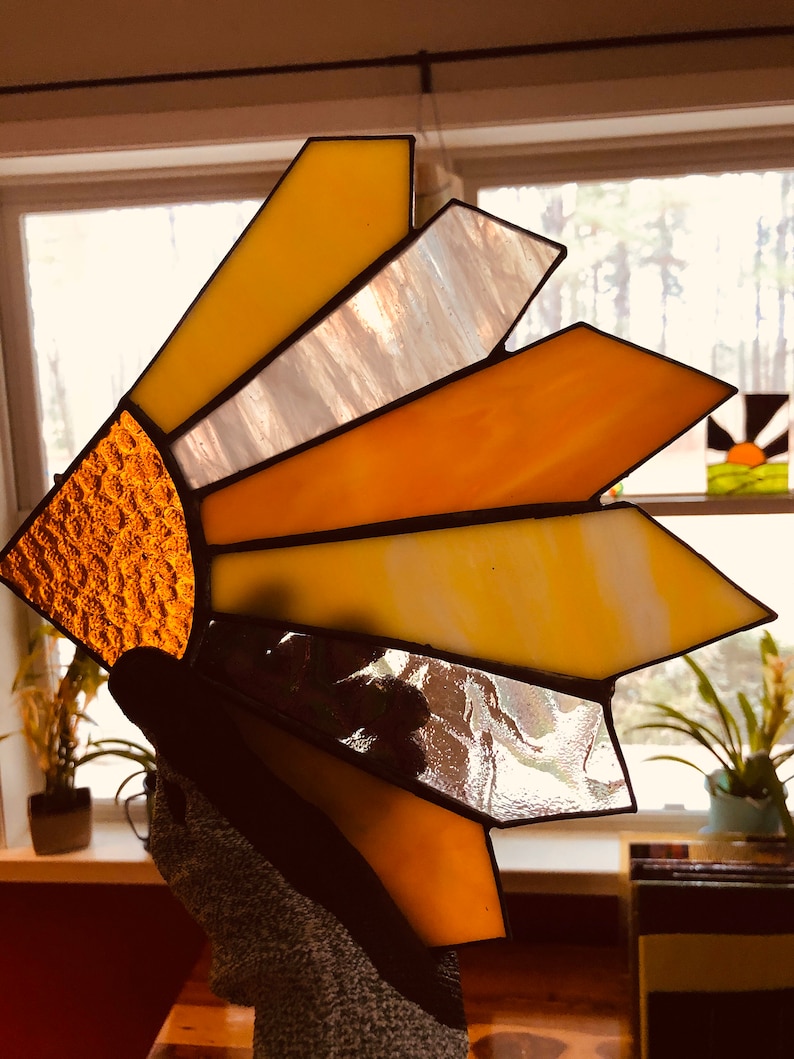 Puede incluir: Un atrapasoles de vidriera con un dise&ntilde;o de sol. El atrapasoles presenta un semic&iacute;rculo naranja texturizado y segmentos radiantes en tonos de amarillo, naranja y blanco. El vidrio est&aacute; enmarcado con soldadura negra.