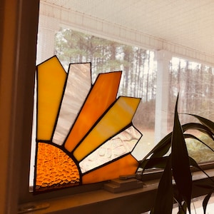 Puede incluir: Atrapasoles de vidriera en una ventana. El dise&ntilde;o presenta un patr&oacute;n de media explosi&oacute;n solar con tonos amarillos, naranjas y blancos. El centro del sol tiene un vidrio texturizado color &aacute;mbar. El atrapasoles est&aacute; montado en un marco de ventana.