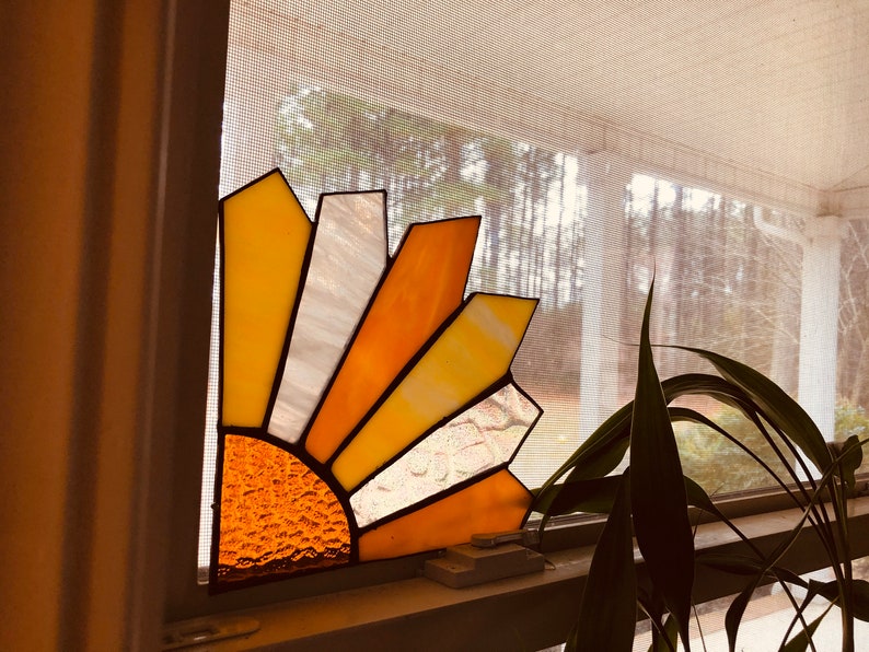 Puede incluir: Atrapasoles de vidriera en una ventana. El dise&ntilde;o presenta un patr&oacute;n de media explosi&oacute;n solar con tonos amarillos, naranjas y blancos. El centro del sol tiene un vidrio texturizado color &aacute;mbar. El atrapasoles est&aacute; montado en un marco de ventana.
