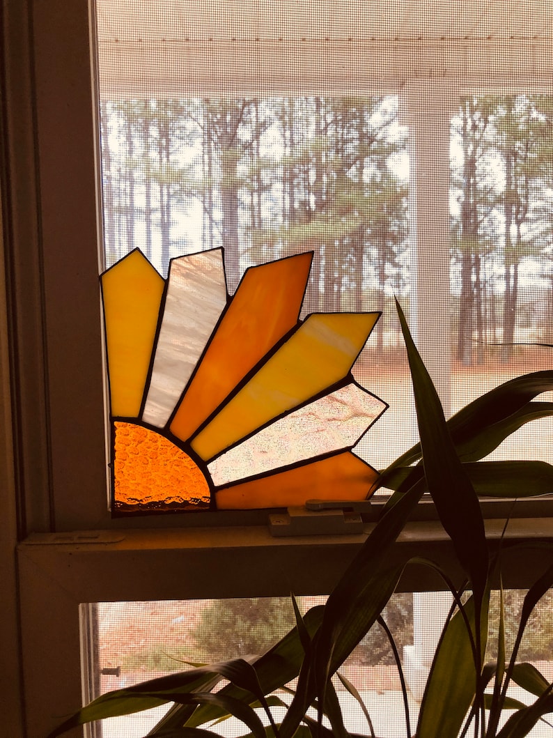 Puede incluir: Un atrapasoles de vidriera con un dise&ntilde;o de medio sol. Presenta segmentos de vidrio amarillo, naranja y blanco, delineados en negro. El atrapasoles est&aacute; colocado en una ventana, con una vista de &aacute;rboles afuera. El dise&ntilde;o evoca un amanecer.