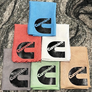 Peut inclure: Plusieurs chiffons de nettoyage en microfibre pliés, de couleurs variées : bleu, rouge, blanc, gris, vert et beige. Chaque chiffon porte le logo noir "Cummins". Les chiffons sont empilés.