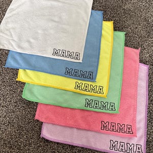 Może przedstawiać: Stos sześciu ściereczek z mikrofibry w różnych kolorach: białym, niebieskim, żółtym, zielonym, różowym i lawendowym. Na każdej ściereczce napis "MAMA" czarnymi literami.