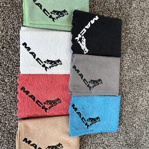 May include: A stack of microfiber cleaning cloths in various colors, including green, white, red, tan, gray, black, and blue. Each cloth features the word "MACK" and a running dog logo in black.