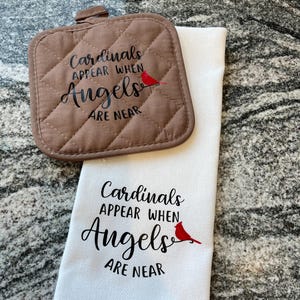 Puede incluir: Una toalla de cocina blanca con el texto "Cardinals appear when Angels are near" y una ilustración de un cardenal rojo. Un posavasos acolchado marrón con el mismo texto y la misma ilustración está encima de la toalla.