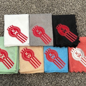 Puede incluir: Una colección de ocho toallas de microfibra en varios colores, cada una bordada con un logotipo rojo y blanco. El logotipo presenta un emblema circular con las letras "KW" y un diseño estilizado.