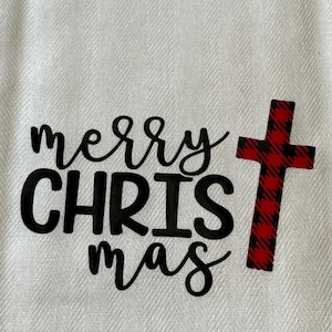 May include: White Christmas-themed towel with the words "merry CHRISTmas" in black script. A red and black plaid cross is on the right side of the towel. The towel has a textured weave.