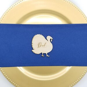 May include: Wooden turkey place card with the word "Dad" written in cursive on a blue napkin and a gold charger plate.