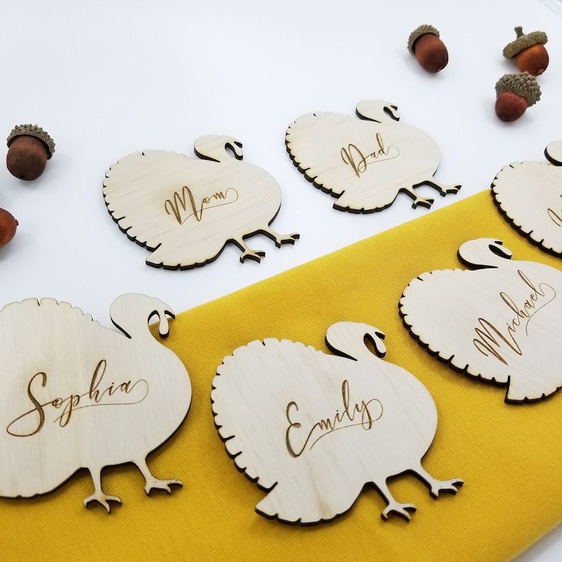 May include: Six wooden turkey place card holders with names written on them. The names are "Sophia", "Emily", "Mom", "Dad", "Michael", and an unreadable name. The place cards are on a yellow tablecloth.