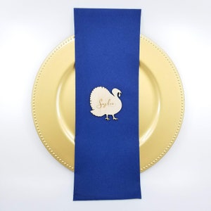 May include: A wooden turkey place card with the name "Sophia" written on it. The place card is sitting on a blue napkin and a gold charger plate.