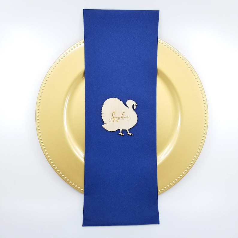 May include: A wooden turkey place card with the name "Sophia" written on it. The place card is sitting on a blue napkin and a gold charger plate.
