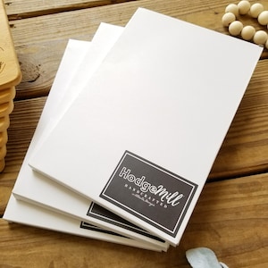May include: Stack of three white blank canvases with a black and white logo that says "Hodgemill Handcrafted - little things"