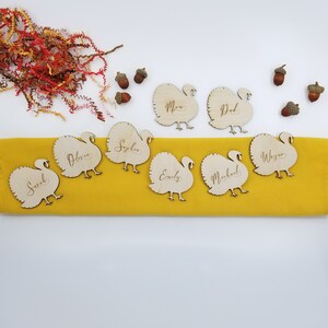 May include: Wooden turkey place card holders with names written on them. The names include Sarah, Olivia, Sophia, Emily, Michael, Wayne, Mom, and Dad. The place cards are on a yellow tablecloth.