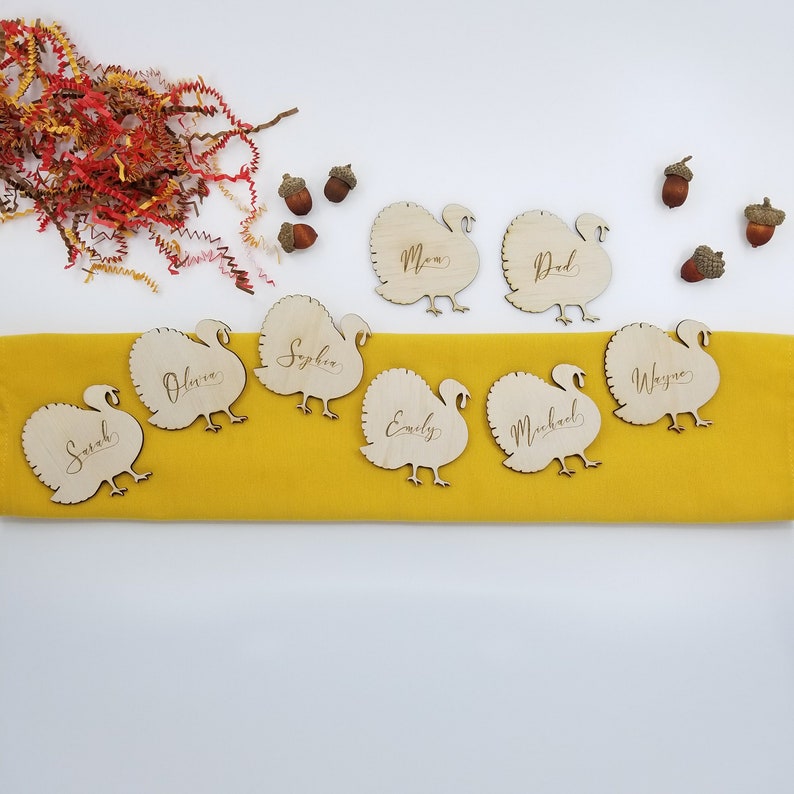 May include: Wooden turkey place card holders with names written on them. The names include Sarah, Olivia, Sophia, Emily, Michael, Wayne, Mom, and Dad. The place cards are on a yellow tablecloth.