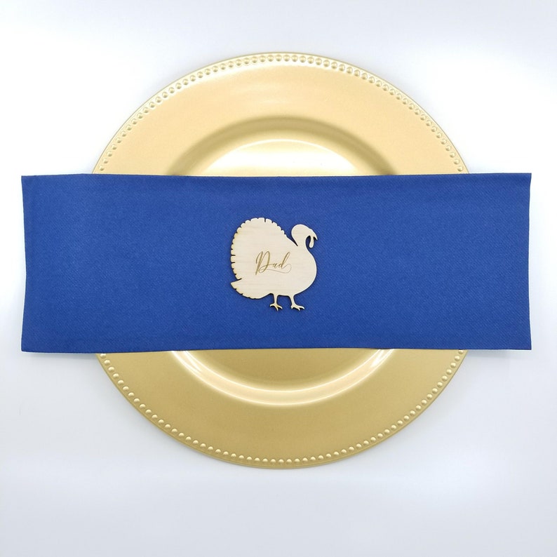 May include: Wooden turkey place card with the word "Dad" written in cursive on a blue napkin and a gold charger plate.