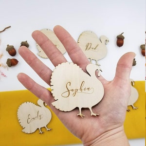 May include: Wooden turkey place card with the name "Sophia" engraved on it. The place card is being held in a hand. There are other place cards in the background with the names "Dad" and "Emily" engraved on them.
