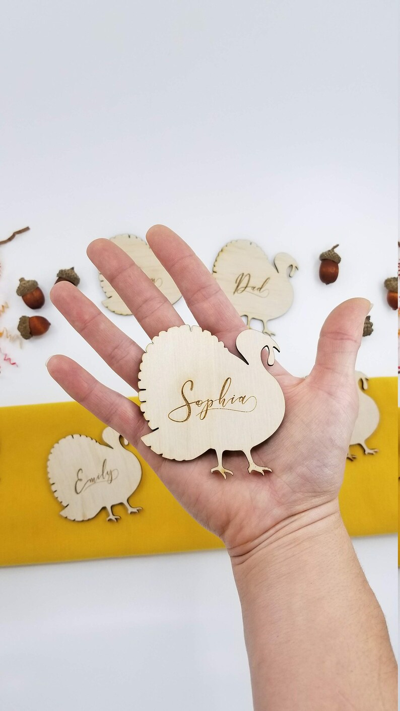 May include: Wooden turkey place card with the name "Sophia" engraved on it. The place card is being held in a hand. There are other place cards in the background with the names "Dad" and "Emily" engraved on them.