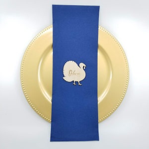 May include: A wooden turkey place card with the name "Olivia" engraved on it. The place card is sitting on a blue napkin and a gold charger plate.