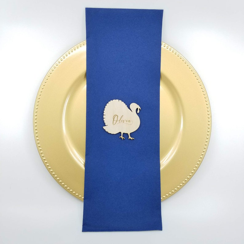May include: A wooden turkey place card with the name "Olivia" engraved on it. The place card is sitting on a blue napkin and a gold charger plate.