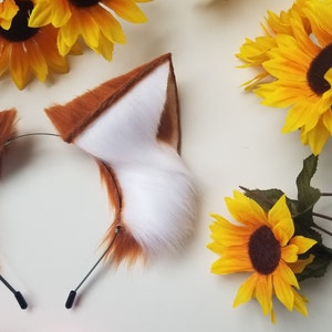 May include: A headband featuring two pointed, faux fur animal ears. The ears have a vibrant orange-brown exterior and a soft, white interior. The headband is constructed from a thin, black metal wire frame with protective black tips at the ends.