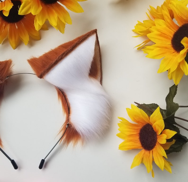 May include: A headband featuring two pointed, faux fur animal ears. The ears have a vibrant orange-brown exterior and a soft, white interior. The headband is constructed from a thin, black metal wire frame with protective black tips at the ends.