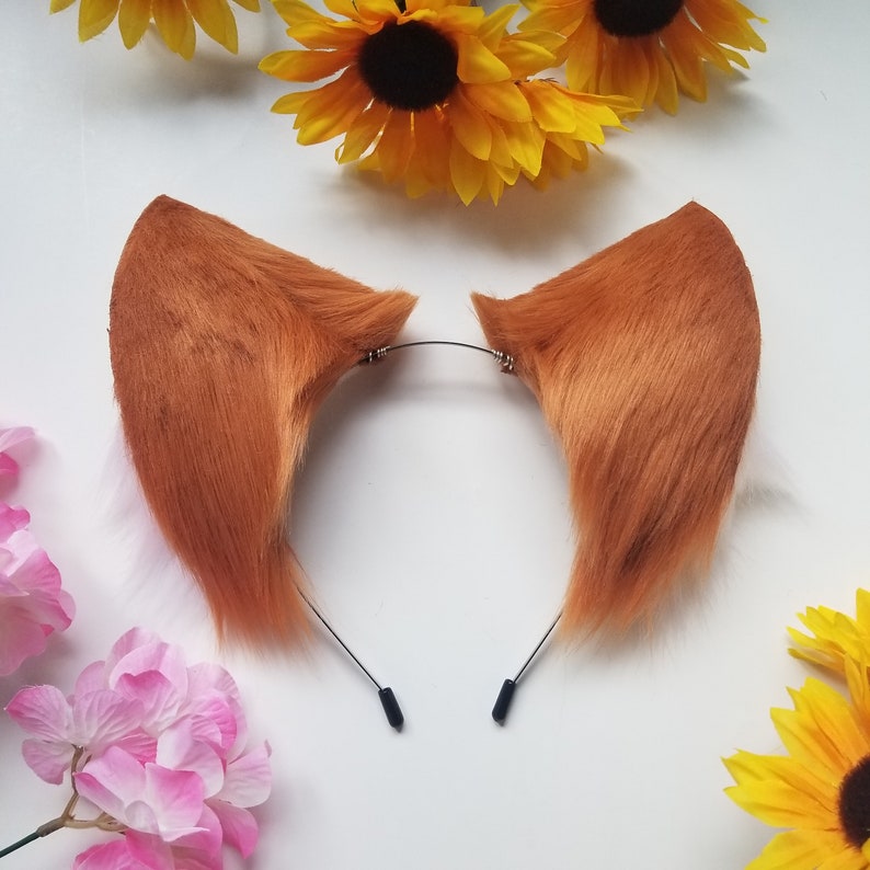 May include: A pair of faux fur fox ears attached to a thin metal headband. The ears are a vibrant orange-brown color with white fur accents visible at the base. The headband is constructed from a slender wire frame with black plastic end caps.