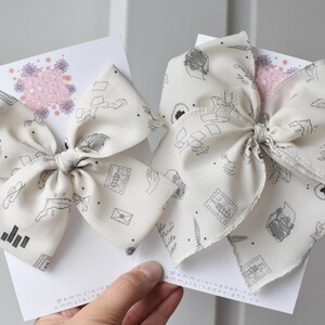 May include: Two white fabric hair bows with a black and white print of a typewriter, envelopes, and other stationery items. The bows are tied with a knot in the center.