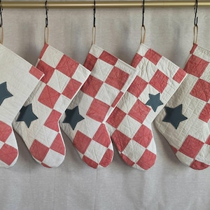 May include: Five quilted Christmas stockings hanging in a row. Each stocking features a red and white checkered pattern with a solid dark gray star appliqué. The stockings are constructed with visible stitching and are suspended by tan loops.