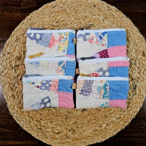 May include: Six small, quilted fabric pouches with zipper closures. The pouches are made from patchwork fabric in various colors, including white, blue, pink, and patterned fabrics. They are arranged on a round, woven mat.