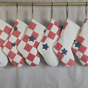 May include: Six quilted stockings hanging from a gold rod, each featuring a red and white checkered pattern. The stockings are decorated with small, dark gray fabric stars appliqued onto the quilted surface, creating a uniform look across all pieces.