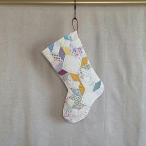 May include: A quilted Christmas stocking constructed from various fabric scraps in a patchwork pattern. The stocking features a mix of floral, checkered, and solid-colored patches in shades of yellow, blue, purple, and white, finished with visible stitching.